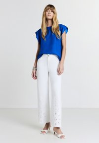 Donna che indossa una blusa corta blu cobalto, jeans bianchi con motivi floreali ritagliati, sandali con tacco bianchi e bracciale d'argento, in piedi.