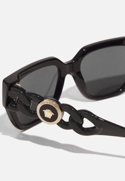 Ochelari de soare negri cu un cadru geometric masiv, lentile întunecate și un emblemă circulară aurie cu un design de cap pe partea cadrului.