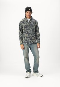 Fleece mikina s maskáčovým vzorem v zelené a béžové barvě, se čtvrtzipovým límcem a malým logem na hrudi, spárovaná s světle modrými džíny.