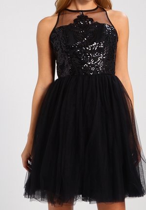 Vrouw draagt een zwarte mouwloze jurk met een lijfje van pailletten in bloemenmotief en een doorschijnende tule rok, staand tegen een effen achtergrond.