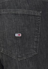 Jeans de mezclilla negra con un pequeño logo bordado con acentos en azul, blanco y rojo en el bolsillo trasero. Primer plano de la costura del bolsillo.