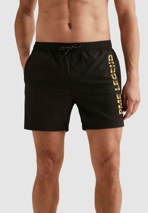 Schwarze Badeshorts mit elastischem Bund und Kordelzug, mit vertikalem gelbem "PME LEGEND" Schriftzug am rechten Oberschenkel, getragen von einem stehenden Mann.