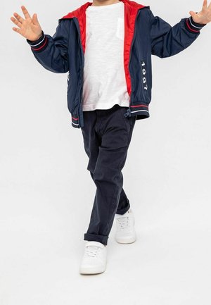 Marineblauwe ritsjack met rode binnenvoering, wit T-shirt, donkere broek en witte sneakers. De jack heeft een jaartalprint en gestreepte accenten.