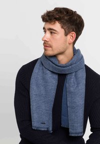 Mand med krøllet brunt hår iført et struktureret blåt tørklæde og en sort ribbet sweater, der ser til venstre mod en ensfarvet grå baggrund.