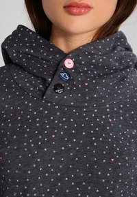 Primo piano della parte inferiore del viso e del collo di una donna che indossa un hoodie scuro a pois con piccoli cuori rosa e tre bottoni colorati sul colletto.
