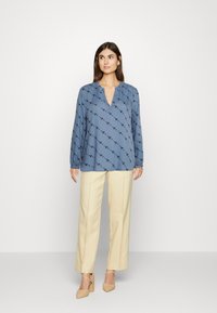Blusa azul de manga longa com decote em V e um padrão preto subtil, combinada com calças de perna larga em tom creme e sapatos de salto neutros.