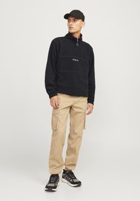 Sudadera negra de vellón con media cremallera y logo blanco, combinada con pantalones cargo beige y zapatos deportivos negros. El sombrero combina con la sudadera.