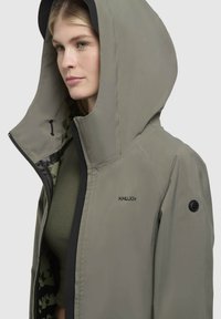 Giacca khaki con cappuccio e dettagli neri, dotata di chiusura con zip e fodera interna decorata. Include un logo sul petto e sulla manica.