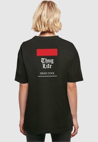 Juodas trumpas rankovė t-shirt, turintis raudoną stačiakampį akcentą ir baltą tekstą: "Thug Life" virš "DEAD COOL" ant nugaros.
