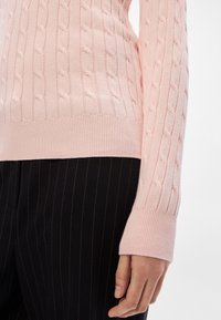 Gros plan d’une personne portant un pull en maille torsadée rose clair et un pantalon noir à fines rayures, avec une main détendue le long du corps.