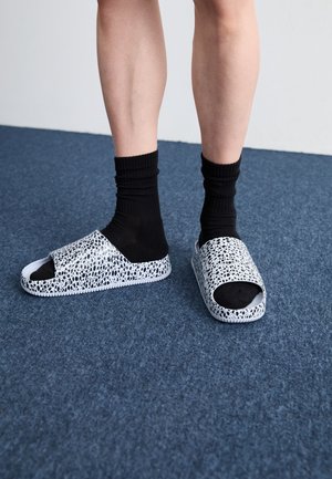 Nogi w czarnych skarpetkach i białych klapkach typu slide z nieregularnym czarnym wzorem kropek, stojące na niebieskim dywanie.