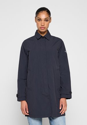 Kobieta ubrana w ciemnogranatowy płaszcz z długim rękawem, z mankietami i kołnierzem zapinanymi na guziki, stojąca na tle jednolitego jasnoszarego tła.