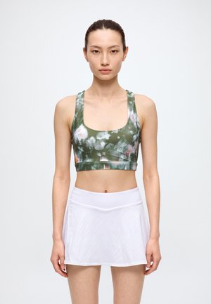 Une femme se tient devant un fond blanc uni, vêtue d’une brassière de sport verte à motifs floraux et d’une jupe de tennis blanche.