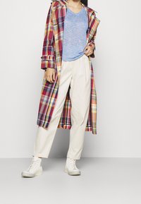 Karierter Trenchcoat in Rot, Gelb und Blau mit Kapuze, getragen über einem hellblauen V-Ausschnitt-T-Shirt und beigen Hosen, abgerundet mit weißen Sneakers.