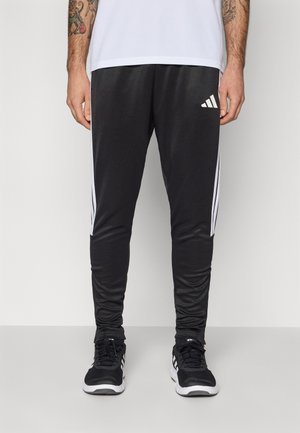 Black athletic pants made of smooth material, featuring white side stripes and an Adidas logo near the waistband. Paired with black sneakers.