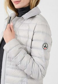 Vrouw in een lichtgrijze gewatteerde jas met het JOTT-logo op de mouw, en daaronder een donkere coltrui.