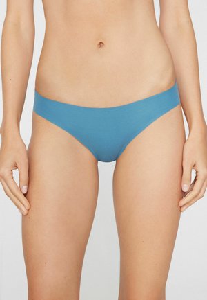 Kobieta nosząca proste niebieskie majtki bikini z niskim stanem, stojąca z rozluźnionymi rękami wzdłuż ciała na jednolitym tle.