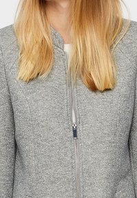 Harmaa vetoketjullinen takki, joka on valmistettu teksturoidusta kankaasta. Takissa on pystykaulus ja etuvetoketju. Yksinkertainen muotoilu ilman näkyviä logoja tai kuvioita.