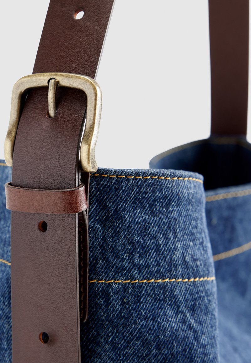 Nahaufnahme einer braunen Leder-Gürtelschnalle, die an blauer Denim-Stoff mit gelber Naht befestigt ist.