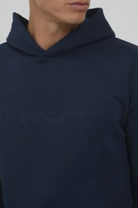 Navy blue cotton hoodie with a textured finish, featuring a drawstring hood and embossed lettering: "BLEND ORIGINAL DENIM EST. 93 - DENMARK."