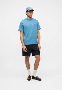 Άνδρας που φορά μπλε καπέλο, μπλε πουκάμισο με κοντά μανίκια, μαύρα σορτς, λευκές κάλτσες και σκουρόκαφέ μοκασίνια, στέκεται μπροστά σε απλό λευκό φόντο.