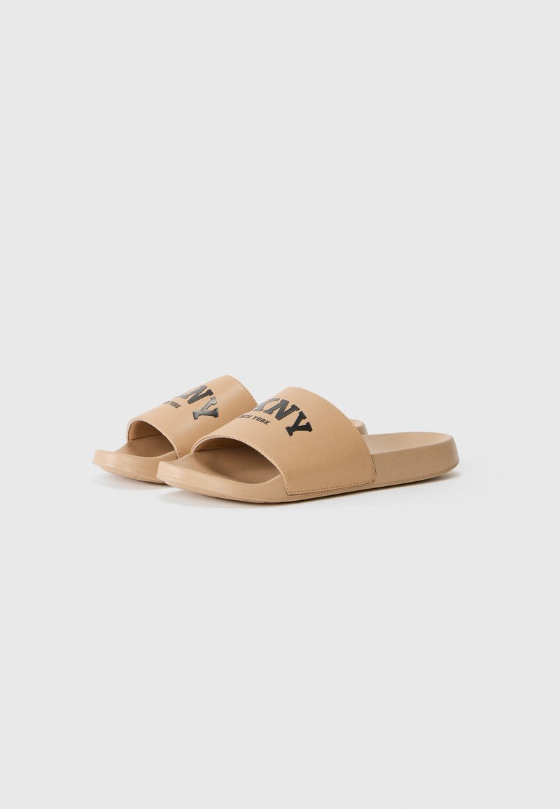Sandalias tipo slide en material sintético color marrón con una amplia tira que presenta el texto "NY" en negro. Suelas planas con un diseño minimalista y textura suave.
