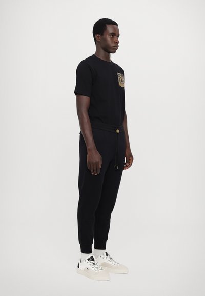 T-shirt en coton noir avec une poche à motifs, associé à un jogging noir avec une taille à cordon de serrage, complété par des baskets blanches.