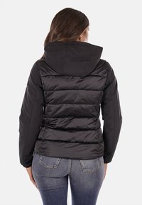 Giacca a vento nera con cappuccio, caratterizzata da un design trapuntato, una texture liscia e polsini elasticizzati. Indossata con jeans blu strappati.