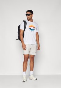 Hvid t-shirt med farverigt bjergmotiv, beige shorts, sort rygsæk og solbriller. Hvide sports sneakers fuldender looket.