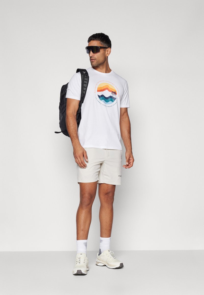 Camiseta blanca con gráfico de montaña colorido, pantalones cortos beige, mochila negra y gafas de sol. Zapatillas deportivas blancas completan el look.