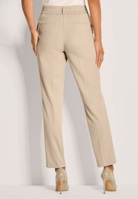 Vrouw in beige rechte broek en bijpassende hogehak schoenen, staande tegen een lichte achtergrond, gezien vanachter.