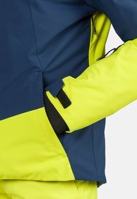Waterbestendige jas met een marineblauwe bovenkant en heldergele onderdelen. Beschikt over een zip pocket en klittenband manchetten voor functionaliteit.