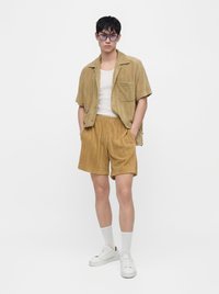 Jeune homme portant une chemise en velours côtelé beige et un short, un débardeur blanc, des chaussettes blanches, des baskets blanches et des lunettes de soleil teintées violettes, debout les mains dans les poches.
