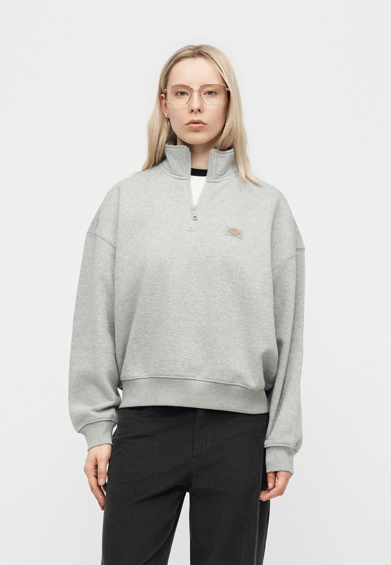 Giovane donna con capelli biondi che indossa una felpa grigia chiara oversize con mezza cerniera e pantaloni neri su uno sfondo bianco uniforme.