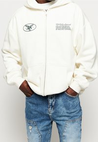 Felpa zip con cappuccio color crema, dotata di tasca a marsupio, con un logo ricamato sul lato sinistro del petto e del testo sul lato destro. Indossata sopra jeans blu strappati.