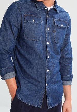 Homme portant une chemise en denim bleue à manches longues avec les poignets retroussés et les poches poitrine boutonnées, debout avec une main dans la poche.
