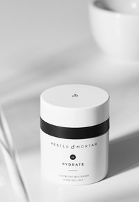 Pot cylindrique blanc avec une bande noire, étiqueté "Pestle & Mortar Hydrate." Le texte inclut le type de produit et une description multilingue. Texture lisse.
