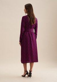 Abito a maniche lunghe in magenta con un motivo geometrico ripetuto. Presenta una vita arricciata ed è abbinato a sandali neri con tacco.