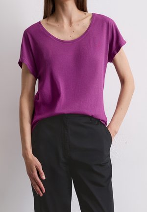 Kvinde iført en lilla top med korte ærmer, der er stoppet ned i højtaljede sorte bukser, stående med venstre hånd i lommen og højre hånd afslappet.