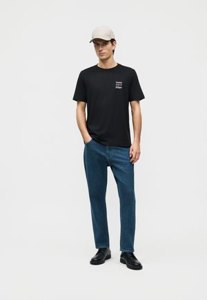 Homme portant une casquette beige, un t-shirt noir Tommy Hilfiger, un jean bleu et des chaussures noires, debout avec une main dans la poche sur un fond blanc.
