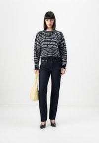 Kvinne iført en marineblå mønstrete genser, mørke straight-leg jeans, sorte hæler og holder en lys gul håndveske, stående mot en hvit bakgrunn.