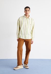 Camisa de manga larga a rayas en amarillo y verde, combinada con pantalones marrones y zapatillas beige/marrón. Tejido suave y corte relajado.