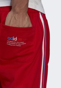 Red athletic shorts with a side pocket, featuring embroidered branding and three vertical stripes in blue, white, and navy along the sides.