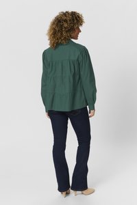 Green, long-sleeve shirt with pleated back detail, paired with dark blue flared jeans and light-colored pointed shoes.