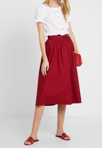 Kobieta ubrana w białą bluzkę z krótkim rękawem, czerwoną spódnicę midi z paskiem, czerwone sandały, trzymająca czerwoną plecioną kopertówkę, stojąca na białym tle.