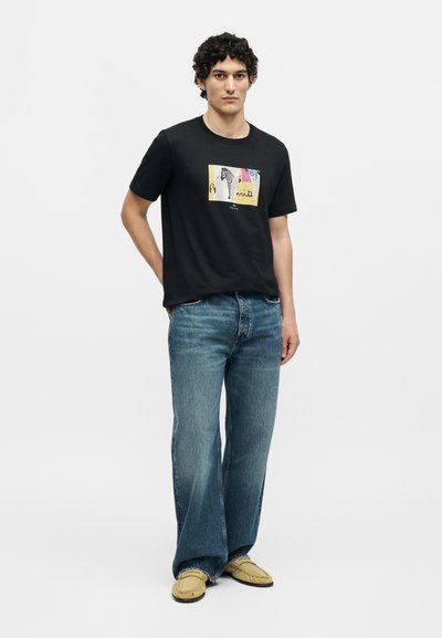 Νέος άνδρας με σγουρά μαύρα μαλλιά, που φοράει μαύρο γραφικό μπλουζάκι, φαρδιά μπλε τζιν και μπεζ μοκασίνια, στέκεται μπροστά σε μία απλή λευκή φόντο.