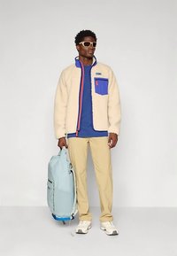 Mees, kes kannab beeži fliisjopet sinise taskuga, sinist särk, beeže pükse, valgeid tossusid, päikeseprille ja hoiab enda kõrval helesinist seljakotti.