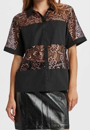 Frau trägt ein kurzärmliges Hemd mit schwarzen Stoffpaneelen und floralen Spitzeneinsätzen, kombiniert mit einem schwarzen glänzenden Lederrock.