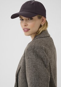 Donkerpaarse baseballpet met een gebogen rand, gecombineerd met een gestructureerde, geruite bruine blazer. De blazer heeft een brede kraag en een relaxte pasvorm.