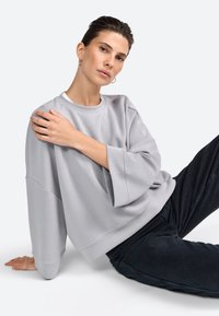 Sudadera de gran tamaño en gris claro con cuello redondo, hombros caídos y mangas largas. Textura suave, combinada con pantalones oscuros.